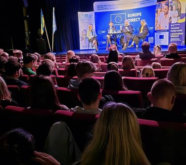 Oświetlona aula. Na fotleach siedzą osoby i słuchają wykładu. Na przodzie na sceenie prelegenci
