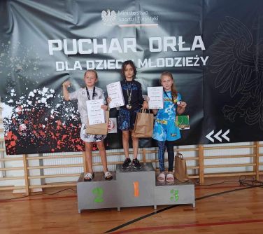 Hala sportowa. Na podium, na tle ciemnego baneru trójka dzieci z dyplomami w dłoni