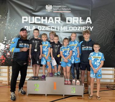 Hala sportowa. Grupa dzieci z trenerem po lewej pozuje na podium