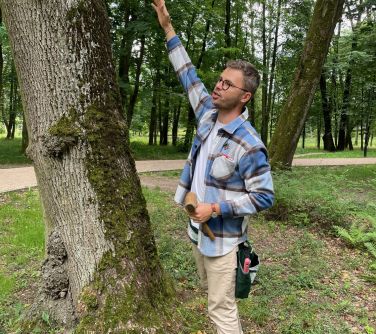 Park w ciągu dnia. Przy dużym drzewie mężczyzna w koszulę w niebieską kratę wskazuje palcem na drzewo