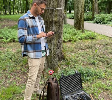 Park w ciągu dnia. Mężczyzna w niebieską koszulę w kratę stoi przy dużym drzewie
