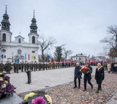 na dworze, dizeń. Trzy osoby z wiązanką maszerują do pomnika. Po lewej biały kościół z dwiema wieżami