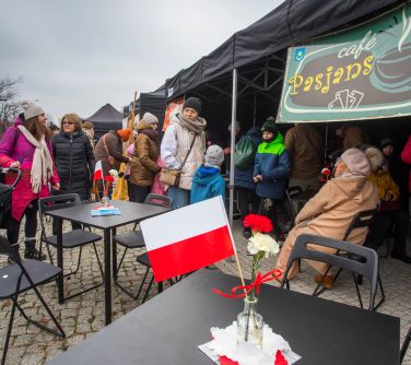 Na dworze, dzień. Przed namiotem czarne kwadratowe stoliki i krzesła. pomiędzy nimi osoby. W tle czarne namioty z tabliczką...
