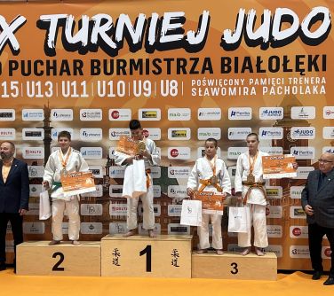 Hala sportowa. Na pomarańczowym tle baneru z logami na podium czworo zawodników w białych kimonach. Po obu stronach mężczyźni