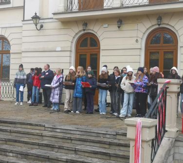 Na dworze w pochmurny dzień. Grupa osób w ubraniach stoi na tarasie przed pałacem
