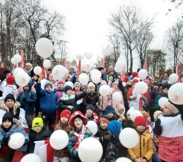 Na dworze w pochmurny dzień. Grupa dzieci w kurtkach i czapkach trzyma białe balony. W tle drzewa bez liści