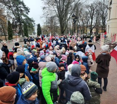 Na dworze w pochmurny dzień. Duża grupa dzieci i dorosłych stoi na placu przed pałącem z flagami biało-czerwonymi. W tle...
