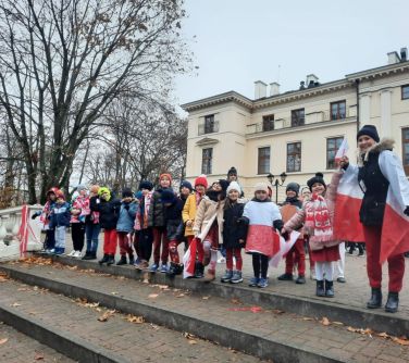Na dworze w pochmurny dzień. Grupka dzieci stoi na górze schodów. Niektórzy trzymają flagi biało-czerwone. Kilkoro ma...