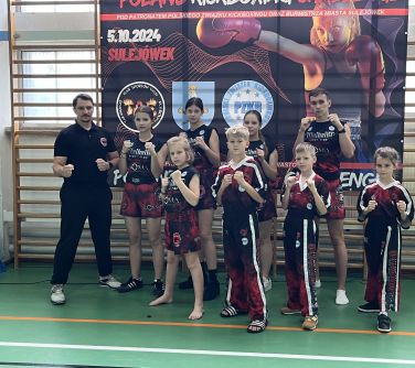 Hala sportowa. Grupa zawodników z trenerem w czarno-czerwonych strojach klubowych pozuje przed drabinkami. Stoją w pozycji...