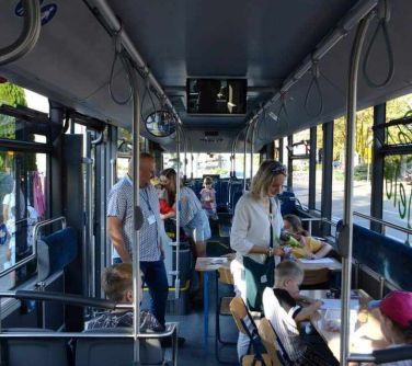 Wnętrze autobusu, dzień. Autobus stoi. Dzieci siedzą przy stole i malują kredkami na kartkach. Dorośli stoją