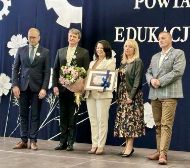 Pomieszczenie. Pięć osób stoi przed granatową kotarą z białymi napisami i kwiatami. Po środku kobieta z bukietem kwiatów i...