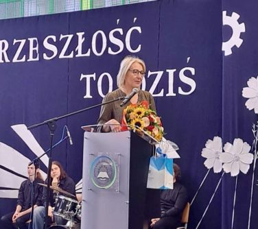 Pomieszczenie. Kobieta w okularach przemawia przy mównicy. Stoi bokiem. Trzyma w ręku bukiet kwiatów i prezent. W tle...