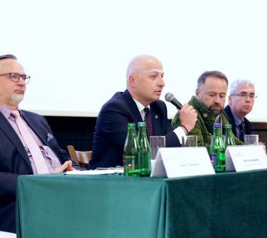 Pomieszczenie. Na tle białej ściany siedzi czterech mężczyzn na krzesłach. Siedza przy stole z zielonym obrusem. Jeden z...
