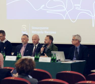 Pomieszczenie. Pięciu mężczyzn siedzi za stołem przykrytym zielonym obrusem. W tle biały ekran z treścią.