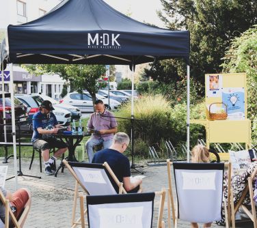 Dwór, w ciągu dnia. Pod namiotem siedzi dwóch mężczyzn przy stole. Czytaja do mikrofonów komiksy. Na przodzie leżaki. na...