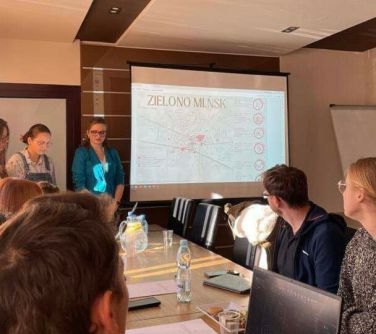 Pomieszczenie. Przy stołach siedzą osoby. na przodzie trzy osoby stoją przy ekranie z wyświetlana treścią
