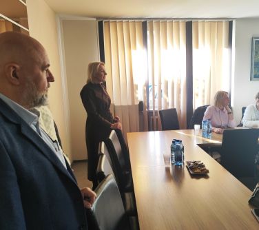 Pomieszczenie, dzień. Kobieta i mężczyzna stoją przed stołem. Kobieta przemawia do siedzących osób. W tle jasne rolety