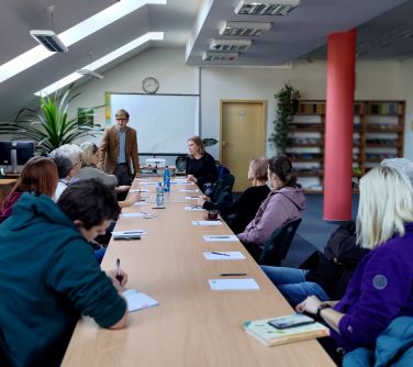 Oświetlona skośna sala. Przy długim stole naokoło siedzą osoby