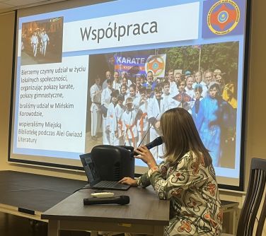 Oświetlona sala. Przy stole kobieta przemawaia do mikrofonu. Patrzy w prawo na treści wyświetlane na ekranie