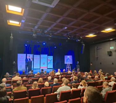 Oświetlona aula. Na fotelach słuchacze. Na scenie banery i osoby