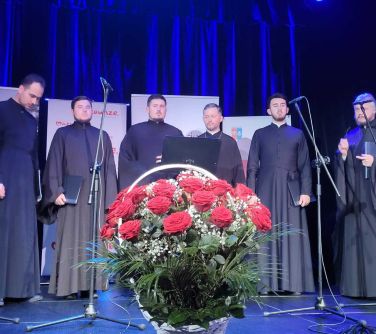 Scena. Sześciu księży w czarnych sutannach na oświetlonej na fioletowo scenie. Na przodzie kosz czerwonych róż