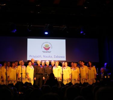 Ciemna sala. Na oświetlonej scenie grupa seniorów. W środku mężczyźni w czarnym ubraniu, czerwonej muszce i poszetce. Po bu...