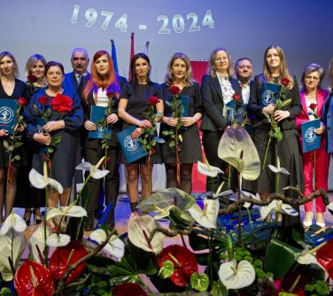 Pomieszczzenie. Grupa osób z długimi, czerwonymi różami w dłoni pozują do zdjęcia. Na przodzie kwiaty