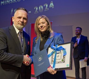 Pomieszczenie. Mężczyzna gratulkuje kobiecie. Kobieta trzyma dyplom i obraz z kokardą