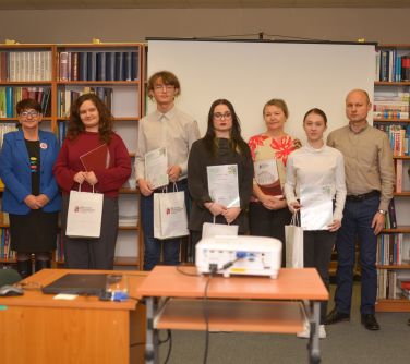 Jasne pomieszczenie. Grupa osób przed półkami pozują do zdjęcia. Nastolatki trzymają dyplomy