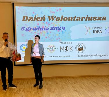 Sala oświetlona. Na ścianie ekran z tytułem głównym Dzień Wolontariatu. Przed ekranem kobieta i mężczyzna który przemawia...
