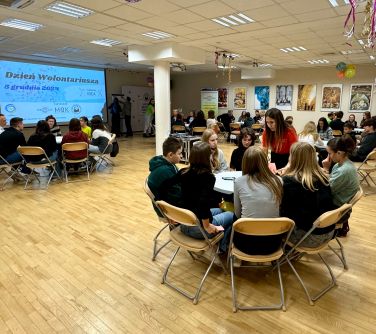 Oświetlona sala konferencyjna. Przy okrągłych stołach na krzesłach siedza osoby. Po lewej duży ekran z tytułem Dzień...