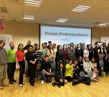 Oświetlona sala konferencyjna. Na tle dużego ekranu z tytułem Dzień Wolontariusza grupa osób