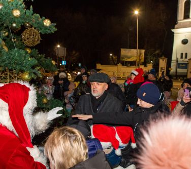 Zmierzch, na dworze. Mikołaj i mężczyzna z grupą dzieci przygotowują się do odpalenia światełek na choince