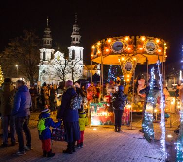 Wieczór, na dworze. Oświetlona karuzela. naokoło ludzie. W tle biały kościół z dwiema wierzami