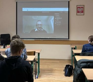 Oświetlona sala lekcyjna. uczniowie w ławkach. Na przodzie na ekranie wyświetlny mężczyzna