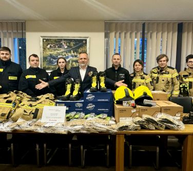 Oświetlony pokój. Na stole ubrania, kaski, buty i akcesoria dla strażaków. Za stołem grupa uradowanych osób