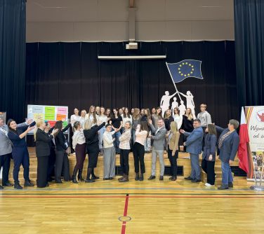 Aula z drewnianą podłogą. Osoby na pierwszym planie gestykulują w kierunku grupy stojącej na podwyższeniu. Wokół sceny...