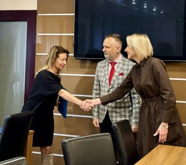 Pomieszczenie. Przy stole trzy osoby. Kobieta gratuluje kobiecie. Po środku mężczyzna