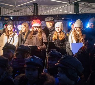 Na zadaszonej scenie grupa osób śpiewa. Ubrani w czapki i kurtki