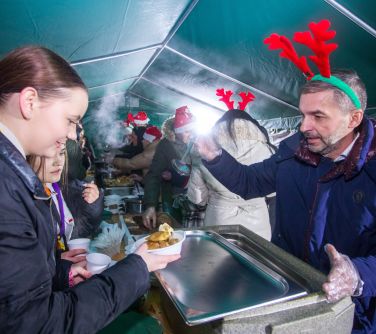 Pod namiotem. Stół z potrawami. Osoby w czapkach mikołaja i rogami reniferów wydaja pożywienie podchodzacym osobom