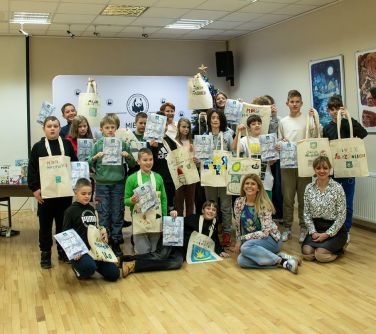 Jasne pomieszczenie. Grupa osób pozuje od zdjęcia. Osoby trzymają prace, tjasne torby lniane w dłoniach. Częścc osób stoi,...