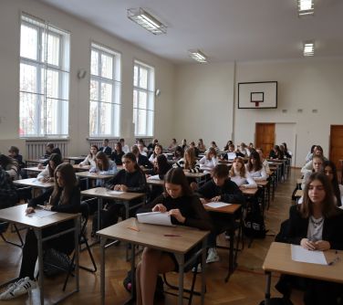 Aula, dzień. Uczniowie piszą egzamin. Siedzą przy pojedynczych ławkach. Sala jest przestronna. W tle widać tablicę do...