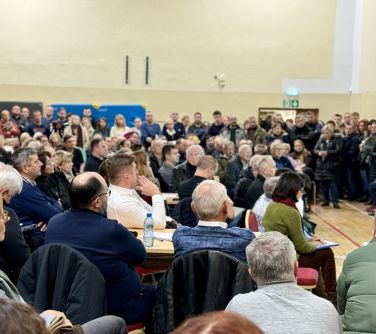 Oświetlona hala. WIele osób. Część siedzi na krzesłach, pozostali stoją. Słuchają