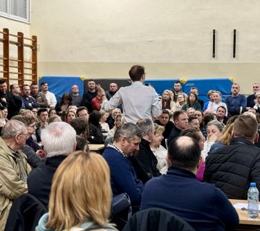 Oświetlona hala. Wiele osób słucha mężczyzne, który przemawia stojąc po środku słuchaczy