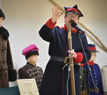 Pomieszczenie. Mężczyzna w stroju historycznym z epoki powstania styczniowego, trzyma broń i przemawia do mikrofonu. W tle...