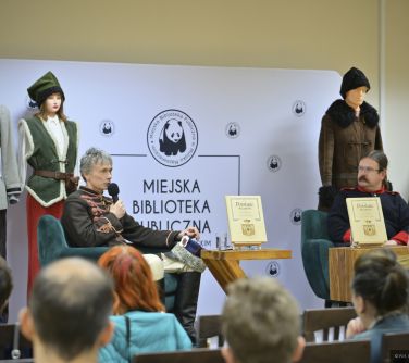 Jasne pomieszczenie. Na fotelach dwie osoby w strojach historycznych. Po lewej osoba przemawia do mikrofonu. W tle biały...