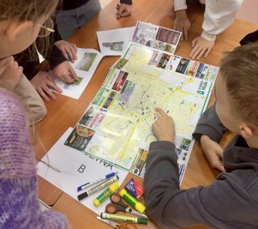 Grupa uczniów pochyla się nad stołem z mapą i materiałami plastycznymi, analizując i pracując wspólnie.