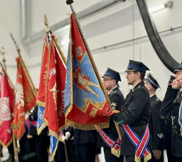Oświetlone pomieszczenie. Pod jasna ściana szpaler strażaków w odświętnych mundurach. Na przodzie cztery pochylone chorągwie