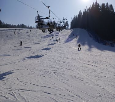 Stok zimą. Dzień. Po śniegu zjeżdżają osoby na nartach i deskach. Po prawej drzewa. Po środku krzesełka kolejki linowej