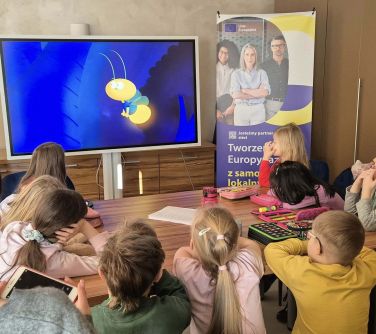 Sala, dzień. Przy stole siedza dzieci. Oglądają wyświetloną kreskówkę, pszczołę. Po prawej beżowe szafy